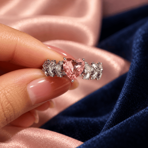 Silver-tone pink heart ring with clear baguette and round stones held between manicured fingers