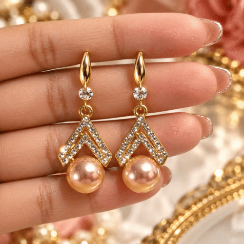 Close-up of a pair of gold-tone double chevron earrings with clear stone accents and peach pearl-look drops held in a hand