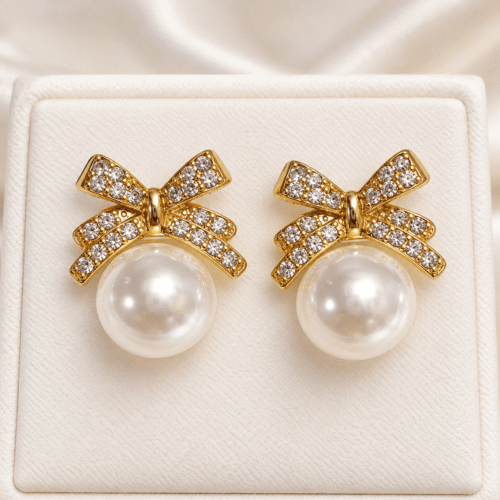 Pair of gold-tone bow earrings with clear stones and white pearl-style beads on a cream stand