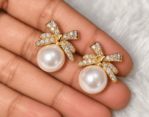 Close-up of gold-tone bow earrings with clear stones and white pearl-style beads held on fingers