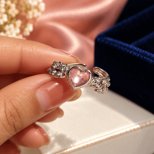 Silver-tone ornate floral ring with a smooth pink heart held between manicured fingers